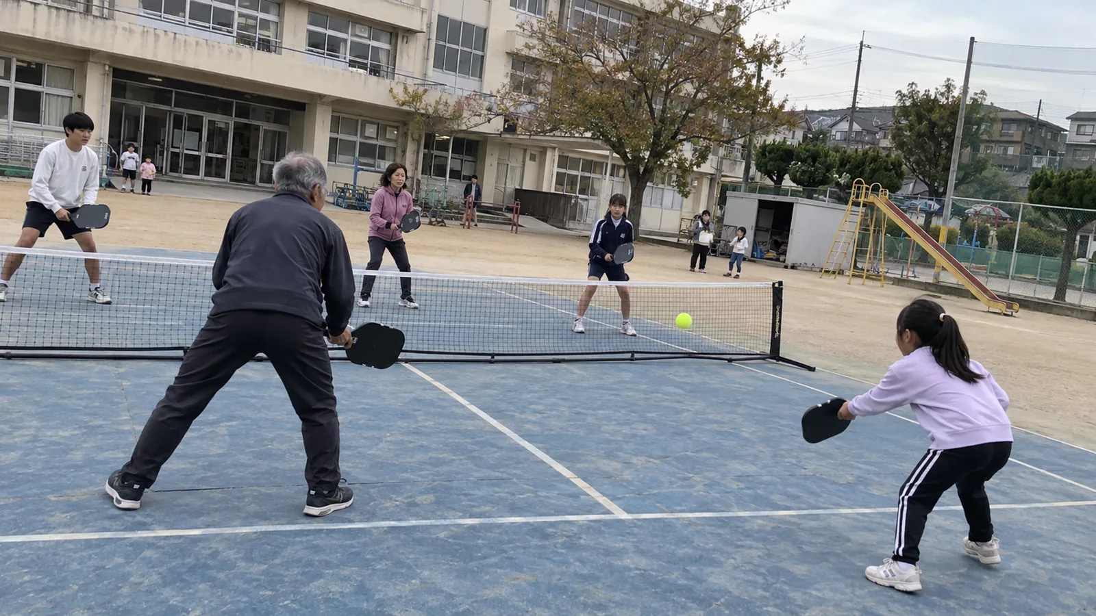 小学校の校庭で、子どもからシニアまでがパドルテニスのダブルスをする様子(イメージ)