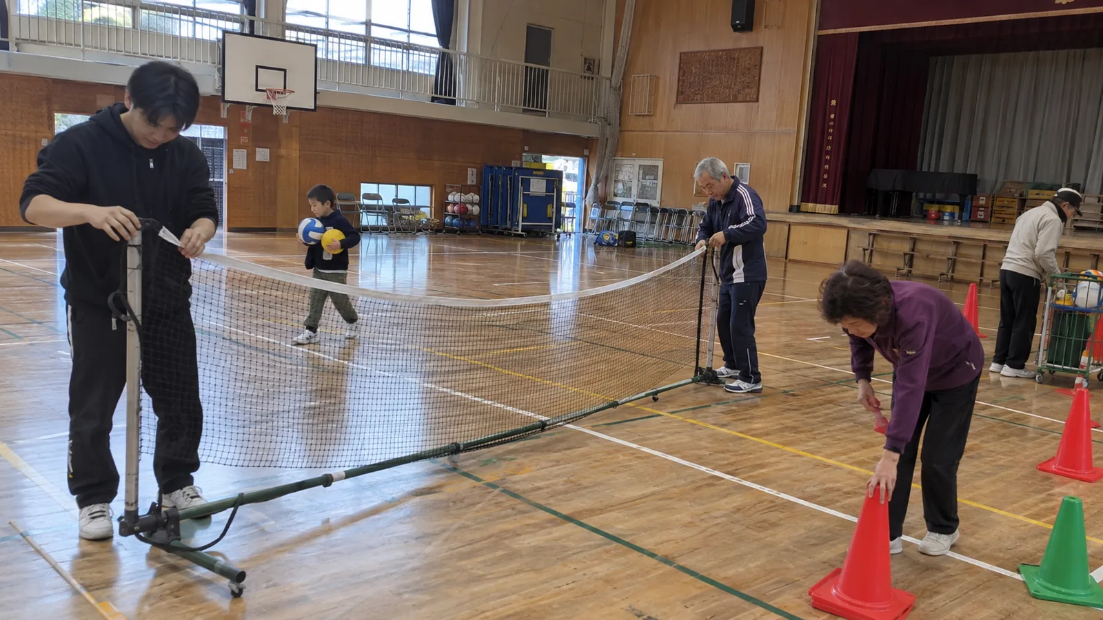 体育館で、大学生とシニアがネットを準備し、子どもがボールを運ぶ様子（イメージ）